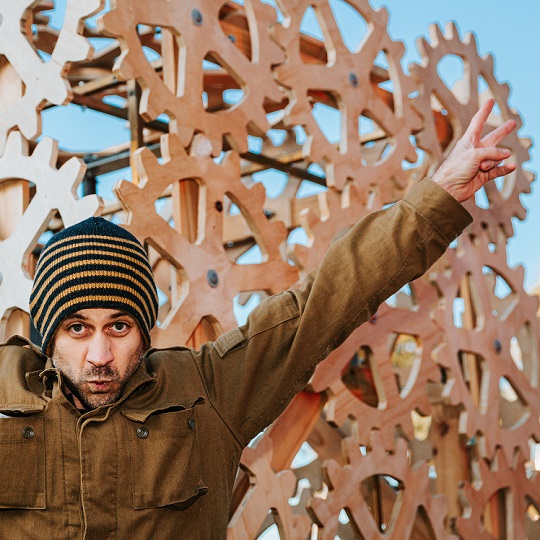 Photo of man with stubble wearing a beanie, he holds his hands up in peace signs on both hands and wears a tan colour jacket, behind him is a wood pine colour cog like sculpture and a blue sky